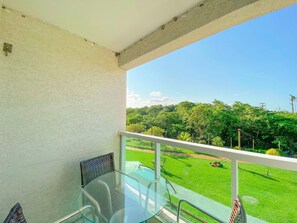 Varanda com mesa e cadeiras de vidro, guarda-corpo branco e vista para um gramado bem cuidado e árvores.