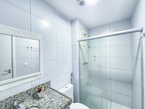 Casa de banho com azulejos brancos, bancada de granito, sanita, cabine de duche com parede de vidro e espelho com moldura decorativa.