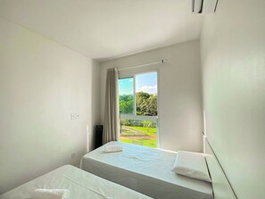 A bedroom with two beds, a window with curtains, and a view of greenery outside.