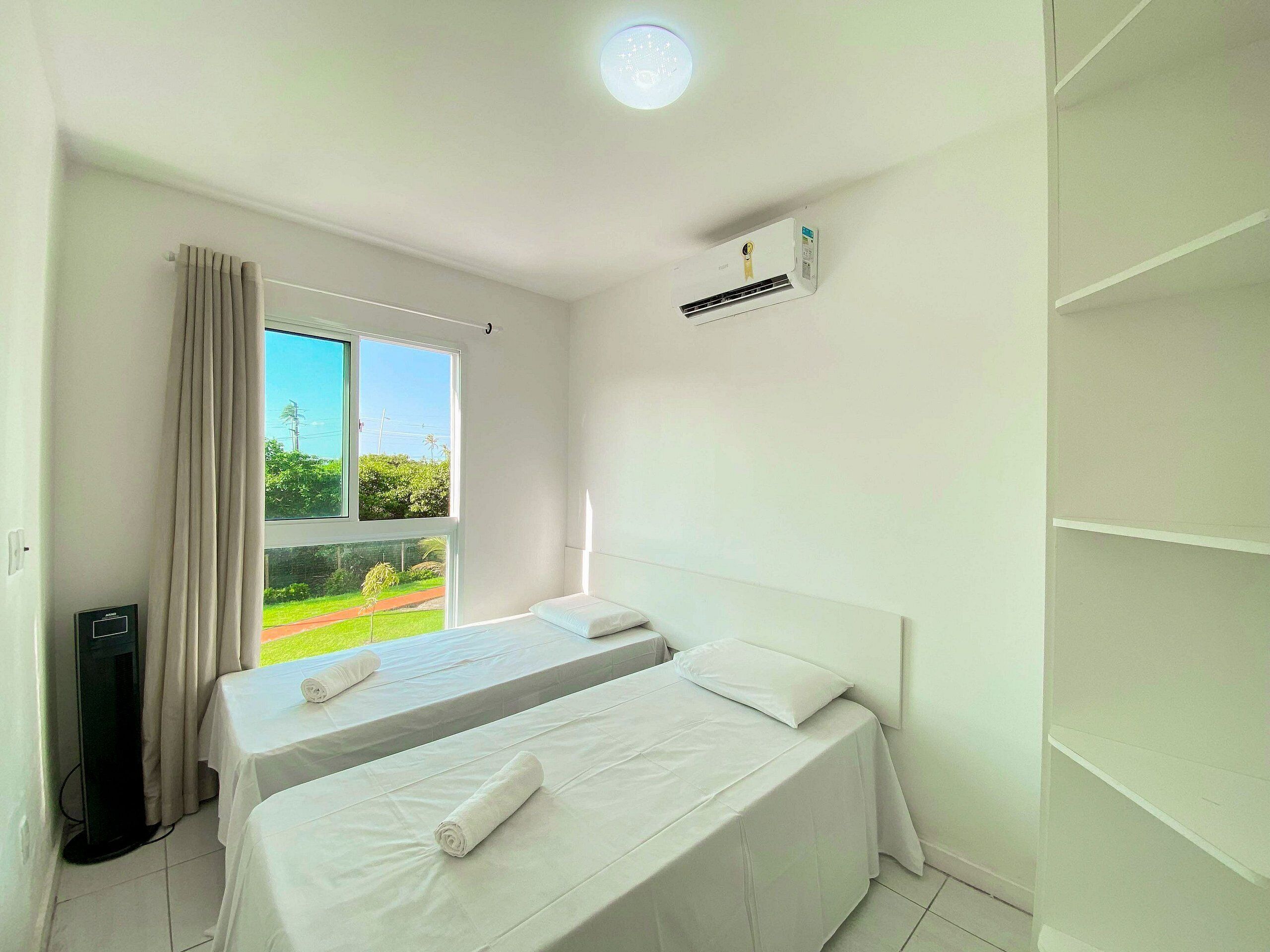 Quarto de hotel com duas camas, ar condicionado, janela com cortinas e vista para o verde lá fora.