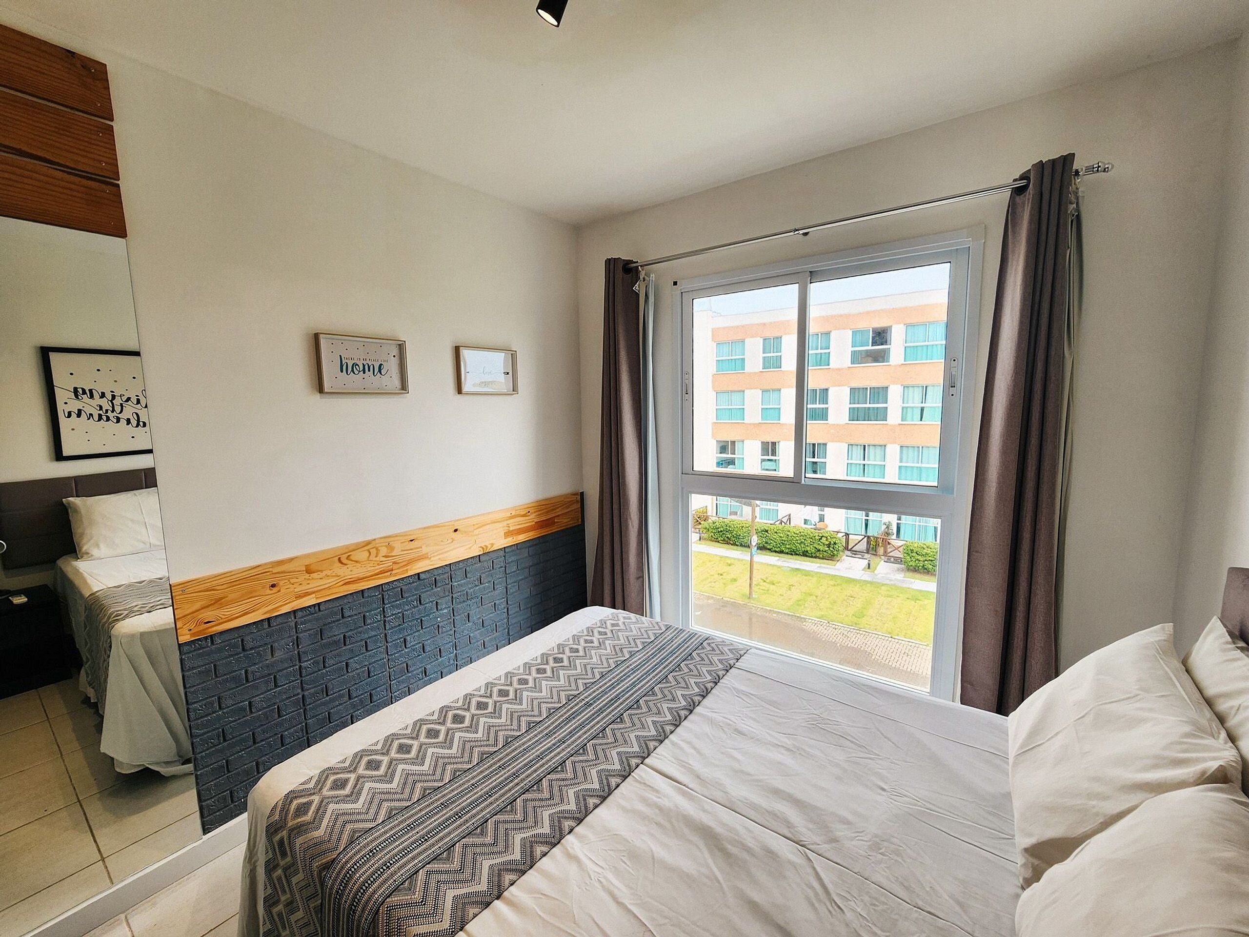 A hotel room with a bed, a window with curtains, a view of a building, and two framed pictures on the wall.