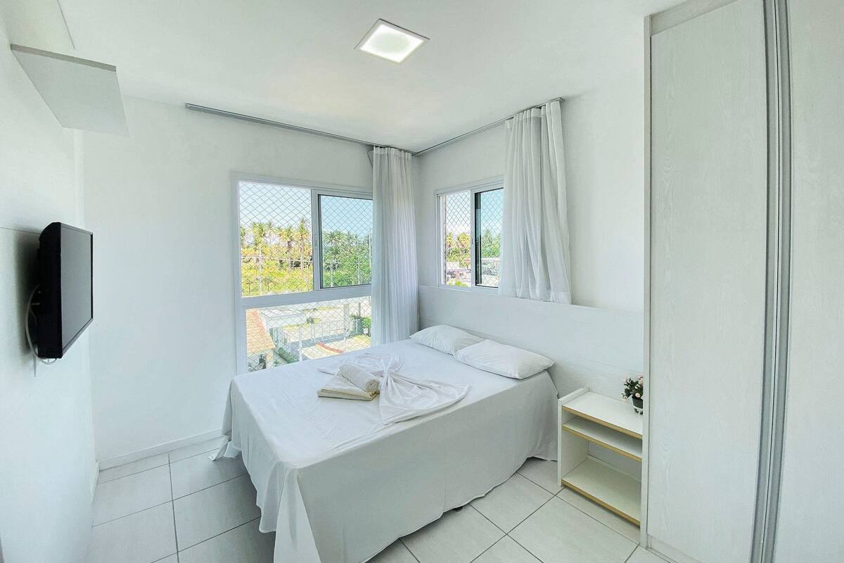 A bedroom with a bed, a bedside table, a TV mounted on the wall, and a window with curtains.