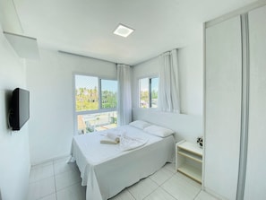 A bedroom with a bed, a bedside table, a TV mounted on the wall, and a window with curtains. - Flats no Muro Alto Condomínio Club (Ipojuca)