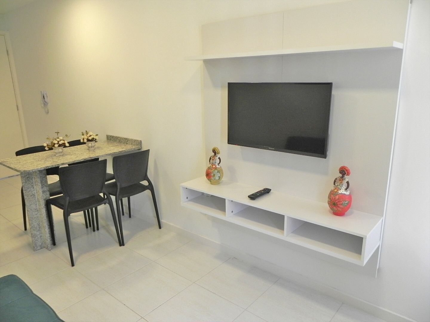 A modern living room with a flat-screen TV mounted on the wall, a dining area with a marble table and chairs, and decorative vases on a shelf below the TV.