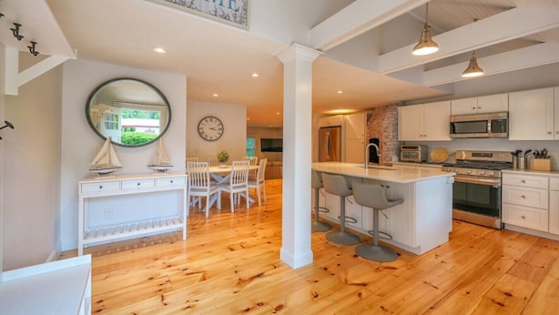 A modern kitchen with white cabinetry, a central island with bar stools, and a dining area with a round table and chairs. - Yarmouth Port Escape Minutes to the Beach (Yarmouth)