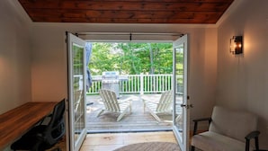 A room with a wooden desk, a chair, a sofa, and a sliding glass door leading to an outdoor deck with two Adirondack chairs and a grill. - Yarmouth Port Escape Minutes to the Beach (Yarmouth)