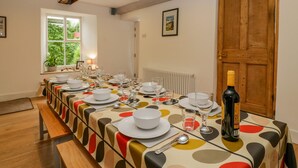 A dining room with a long table set for a meal, a wooden door, and a window with a view of greenery. - Pentre Court Cottage (Abergavenny)