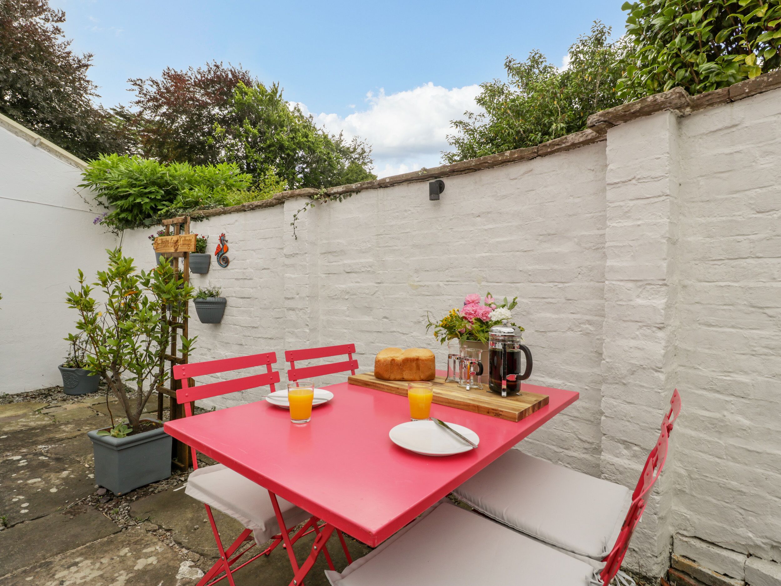 A red table with white cushions, a loaf of bread, a pitcher of juice, and a flower vase in a garden setting.