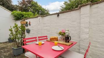 A red table with white cushions, a loaf of bread, a pitcher of juice, and a flower vase in a garden setting.