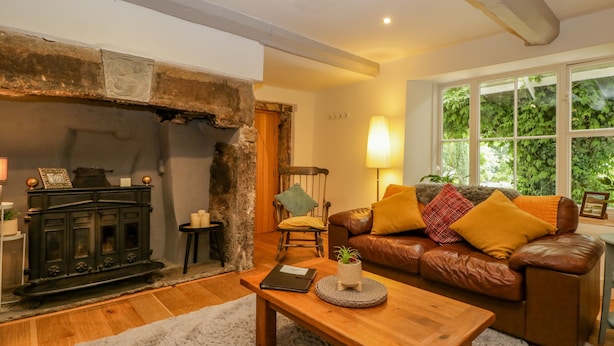 A cozy living room with a fireplace, a leather sofa, a wooden coffee table, and a large window with a view of greenery. - Pentre Court Cottage (Abergavenny)