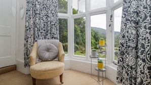 A corner room with a bay window, a chair, a small table with potted plants, and patterned curtains. - Pentre Court Cottage (Abergavenny)