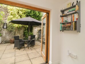 Un patio abierto con una mesa y sillas bajo un paraguas grande, una estantería con libros y un muro de piedra al fondo.