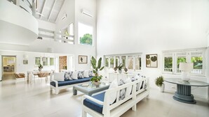 A spacious, well-lit living area with a high ceiling, white furniture, and large windows offering views of greenery. - Closest villa in Casa to Minitas Beach with private pool, garden, screened patio (La Romana)