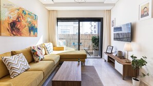 Een moderne woonkamer met een bank, salontafel en televisie. Er is een balkon met uitzicht op gebouwen.
