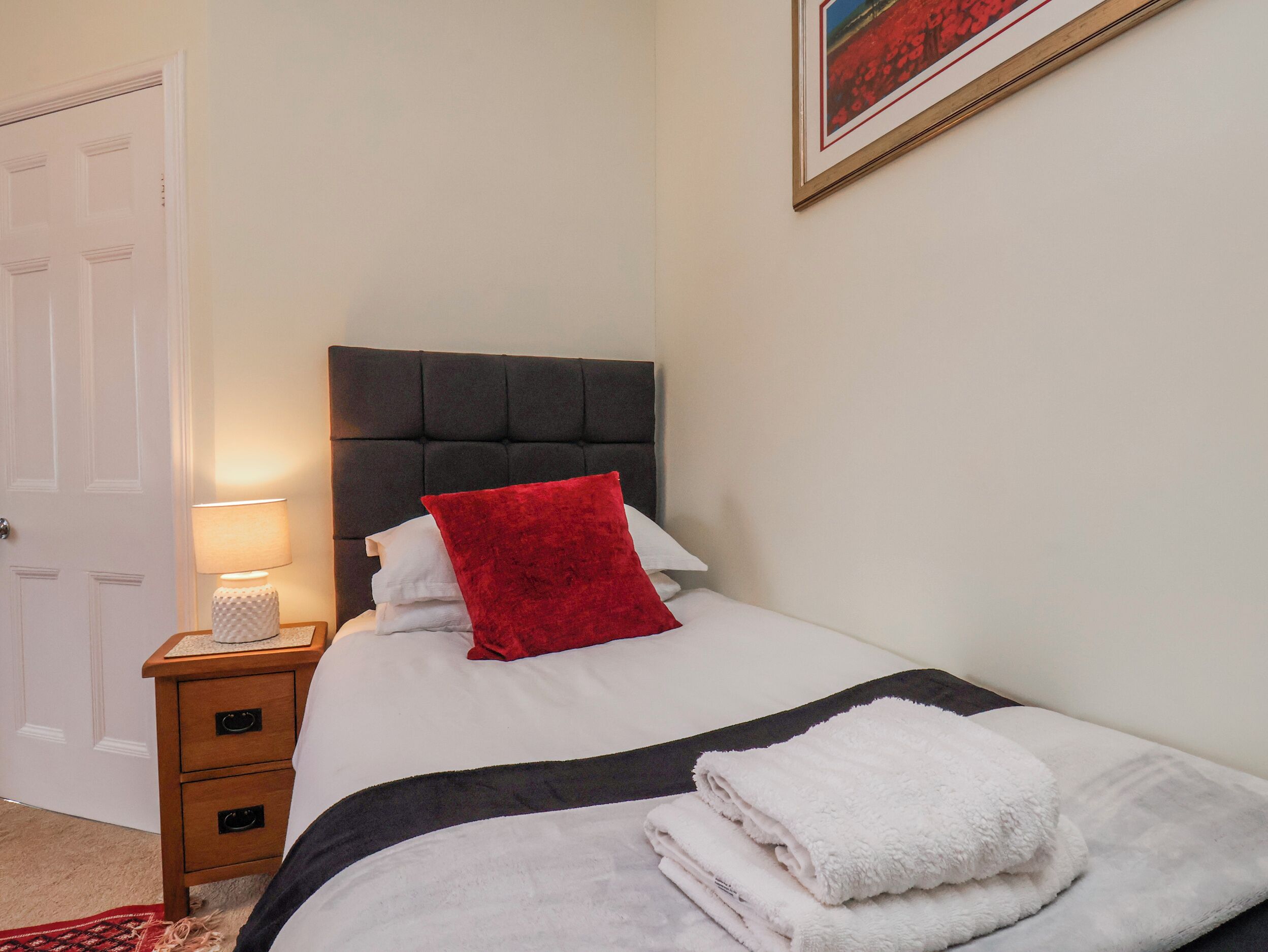 A neatly made bed with a red pillow, a bedside lamp, and a small wooden nightstand with drawers.