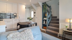 A modern kitchen with white cabinets, a dining table with chairs, a staircase with blue railings, and a living area with a blue sofa.