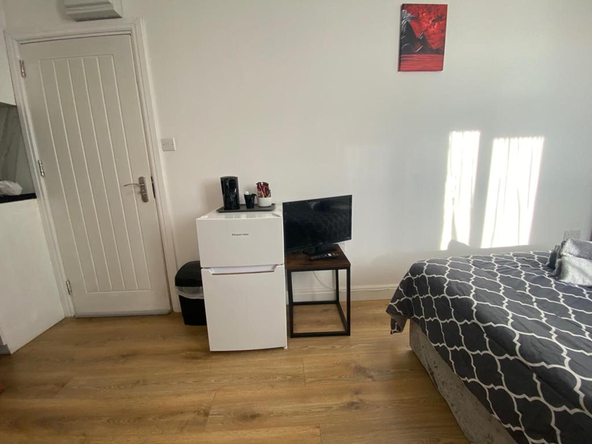 A bedroom with a bed, a television on a stand, a small white refrigerator, and a door.