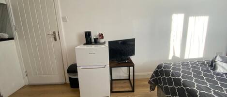 A bedroom with a bed, a television on a stand, a small white refrigerator, and a door.
