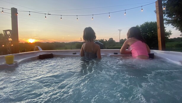 Two people in a hot tub with a sunset view, string lights above, and a clear sky. - Thunderbird Farm - Luxury 5-Bedroom Farmhouse w/ Pool & Hands-On Farm Experience (Kawartha Lakes)