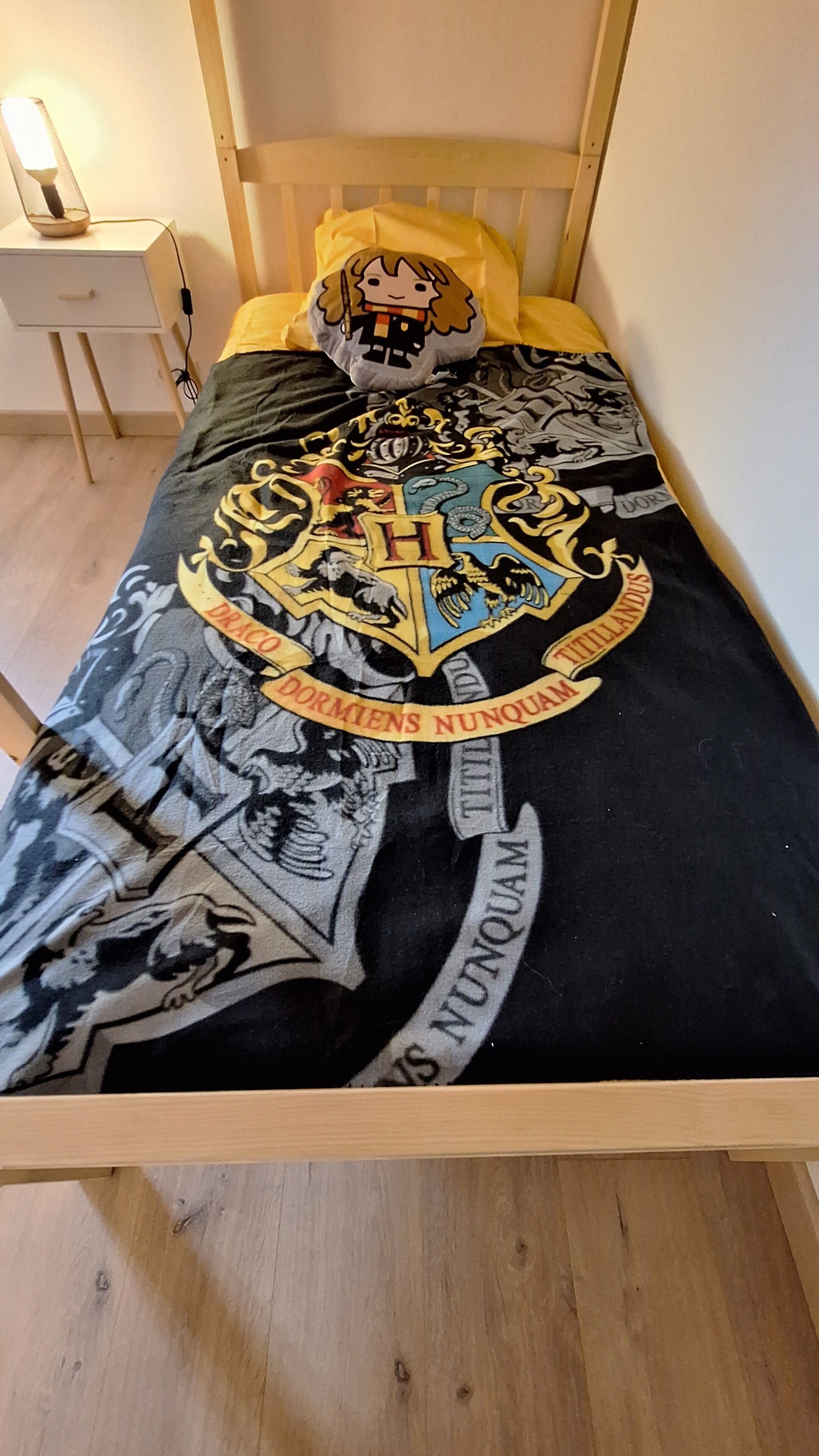 Un lit recouvert d’une couverture noire et dorée ornée d’un blason et de l’inscription « Draco Dormiens Nunquam Titillandus ».