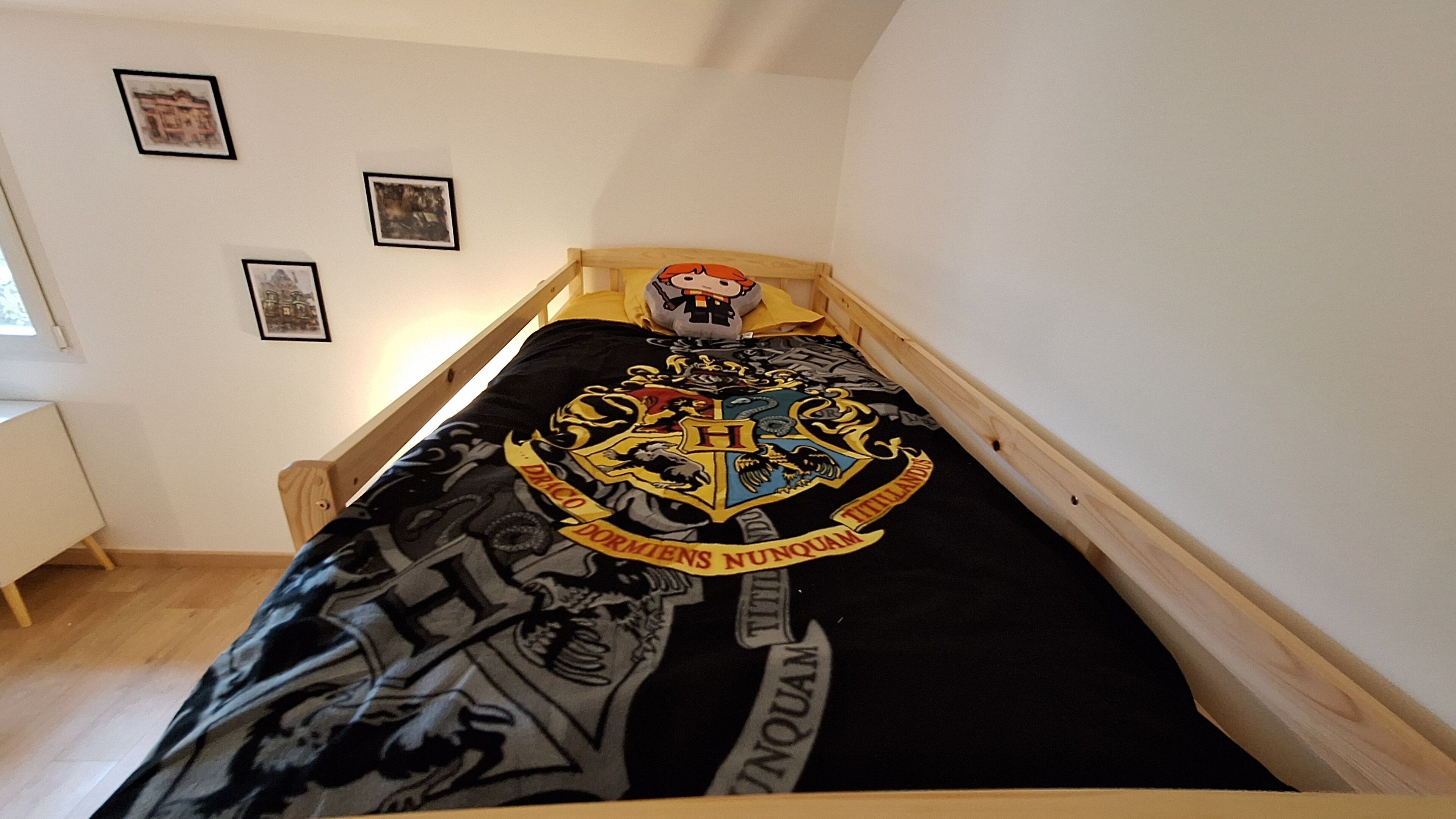 Ein Bett im Studentenwohnheim-Stil mit Hogwarts-Bettwäsche, einem Holz-Kopfbrett und einer weißen Wand mit gerahmten Bildern.