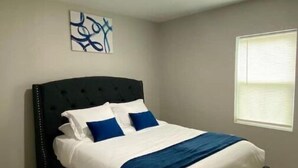 Une chambre à coucher avec un lit recouvert d’un tissu noir, une literie blanche et bleue, un plaid bleu et une œuvre d’art accrochée au mur.