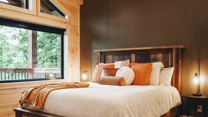 A wooden cabin bedroom with a large bed, a nightstand, a window with a view of trees, and a wooden ceiling.