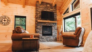 A cozy log cabin interior with a stone fireplace, leather sofas, and large windows.