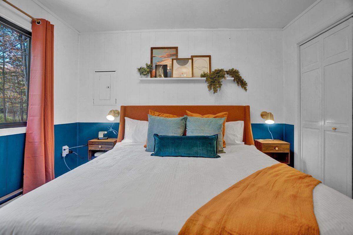 A bedroom with a bed, two bedside tables, a nightstand, a window with curtains, and a wall-mounted shelf with decorative items.