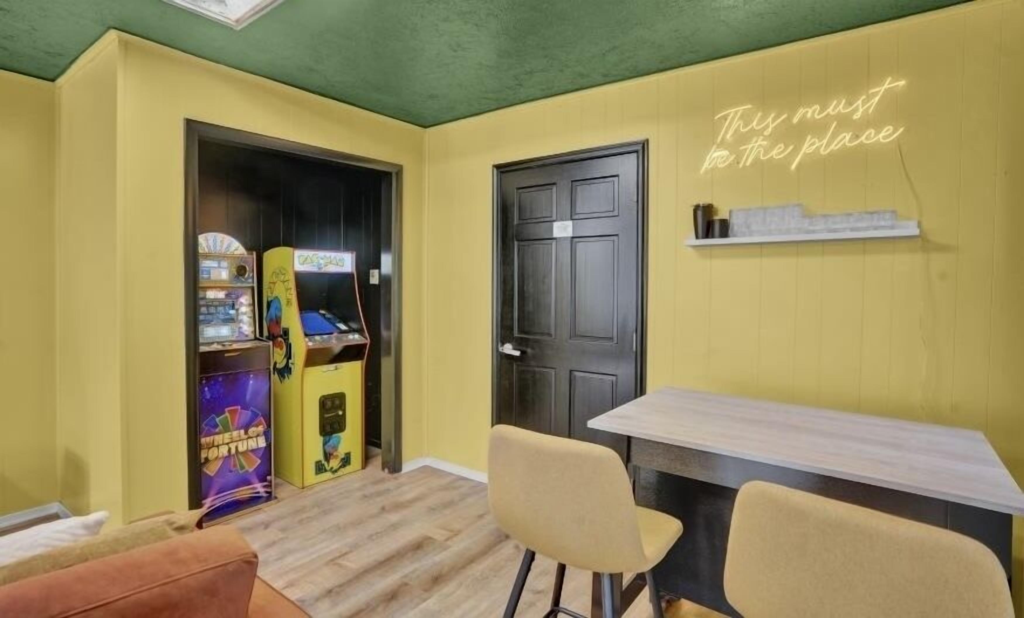 Una habitación con paredes amarillas, una puerta negra, un letrero de neón, una mesa y dos sillas.