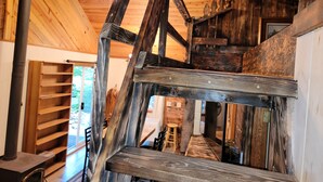 Et hyggeligt sommerhusinteriør med en trætrappe, en loftsrum og en pejs.