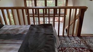 A loft bedroom with a sloped ceiling, a large window, a wooden railing, and a bed with a striped blanket.