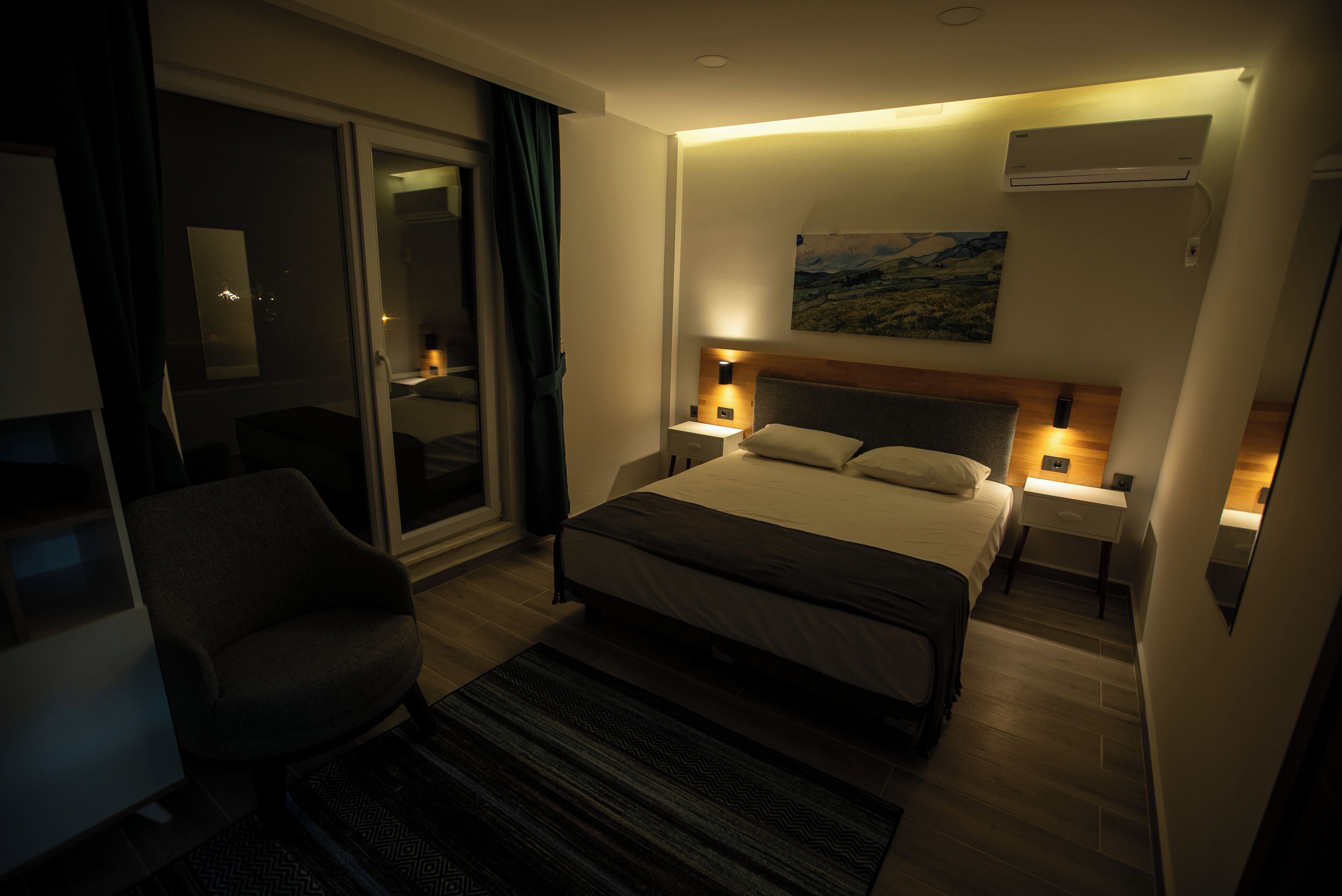A hotel room with a bed, bedside tables, a chair, a large window, and a painting on the wall.