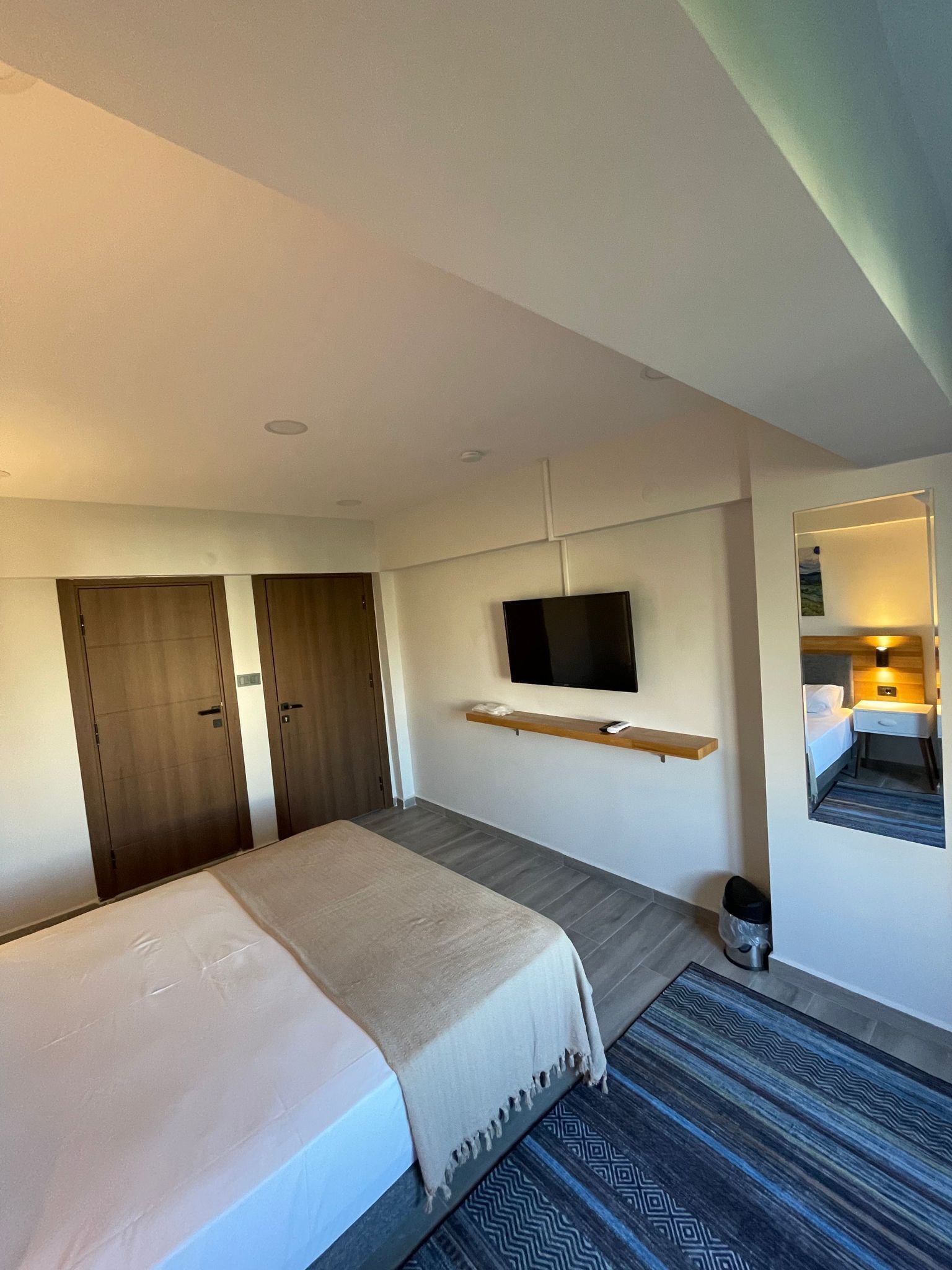 A hotel room with a bed, a TV mounted on the wall, a small bedside table, and a mirror.