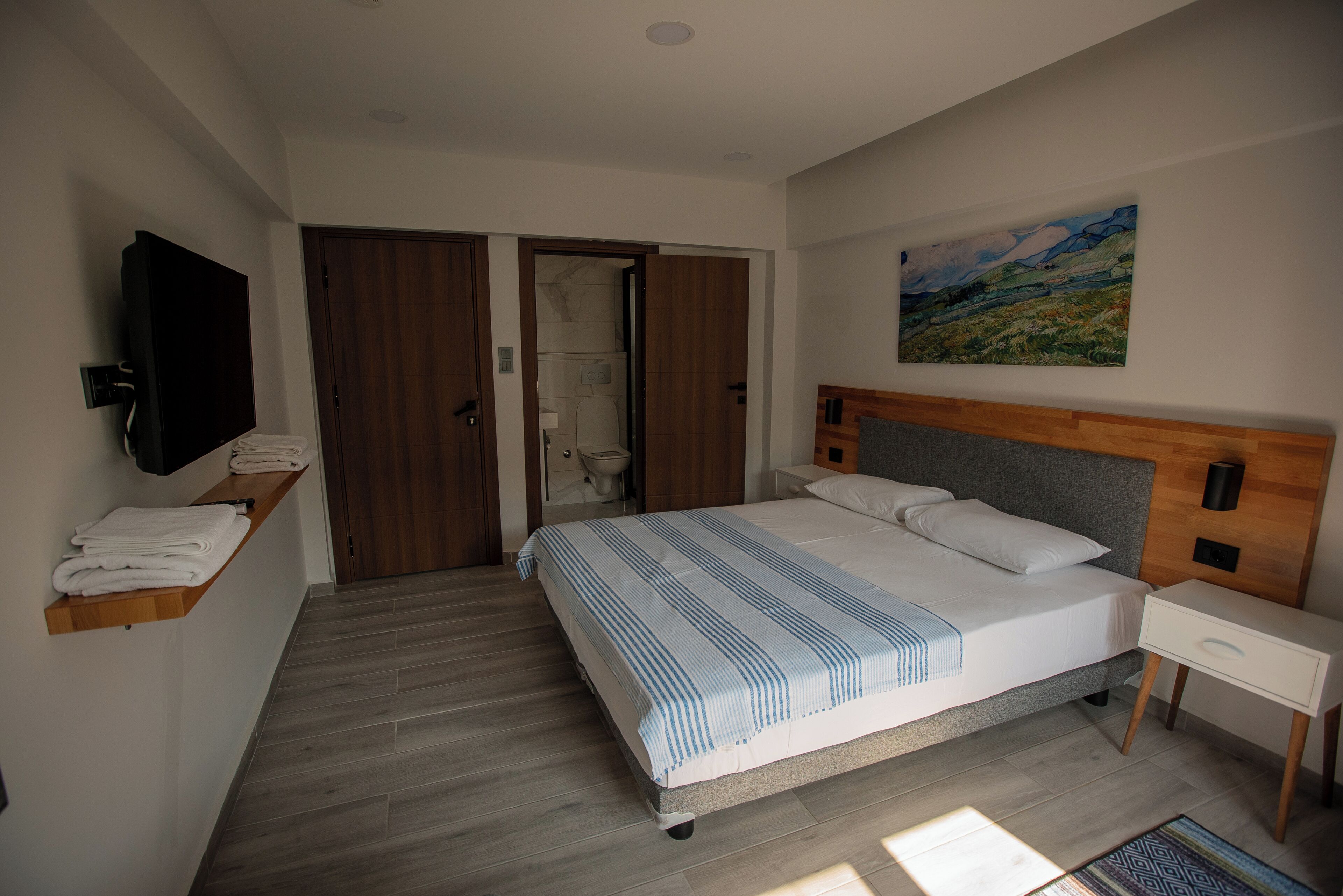 Un dormitorio moderno con una cama grande, un cabecero de madera, una mesita de noche, un televisor montado en la pared y un cuadro sobre la cama.