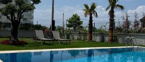 Una piscina con agua azul cristalina, rodeada de césped, palmeras y un edificio al fondo.