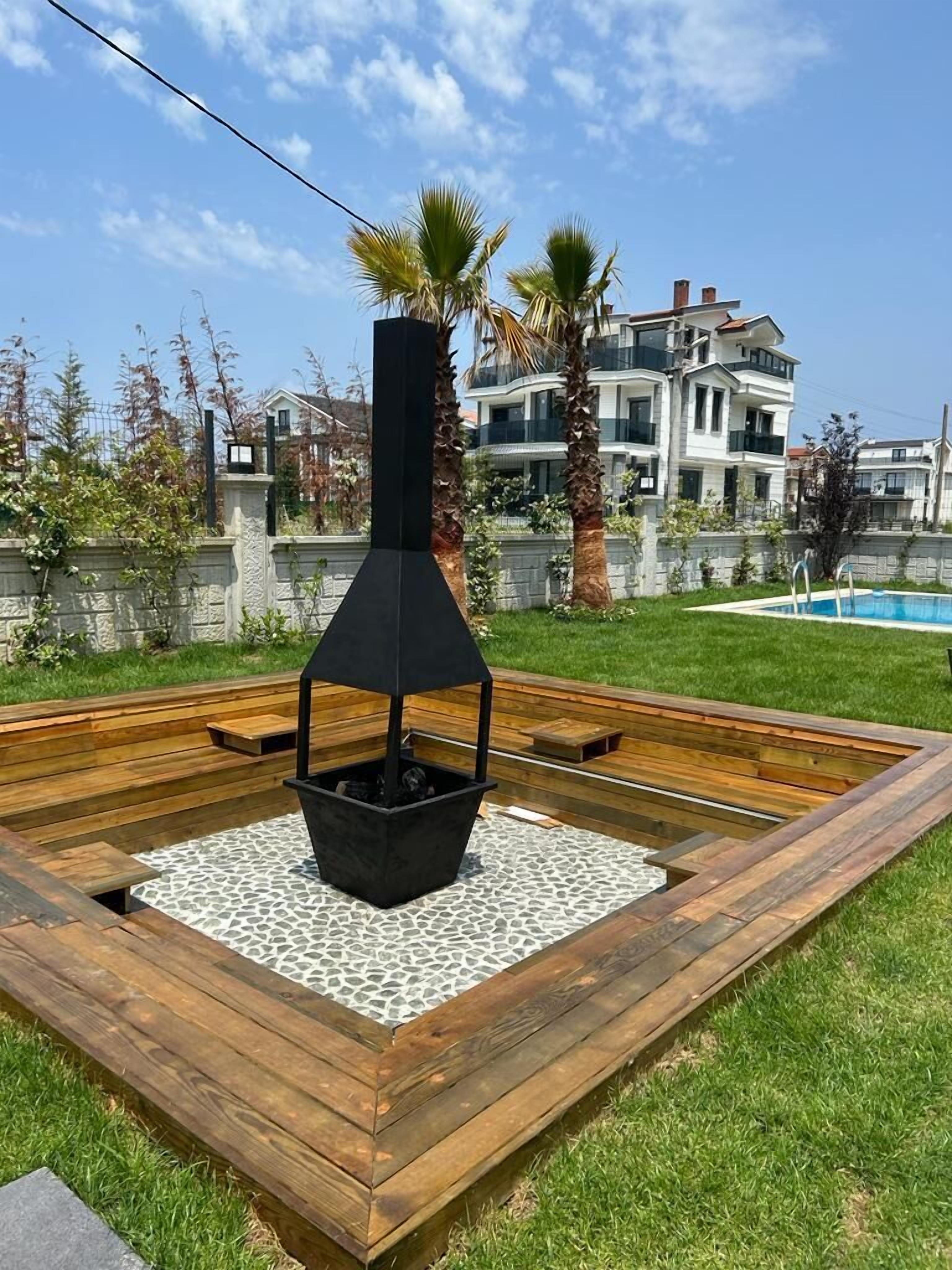 A backyard with a built-in fire pit, a wooden deck, and a swimming pool in the background.