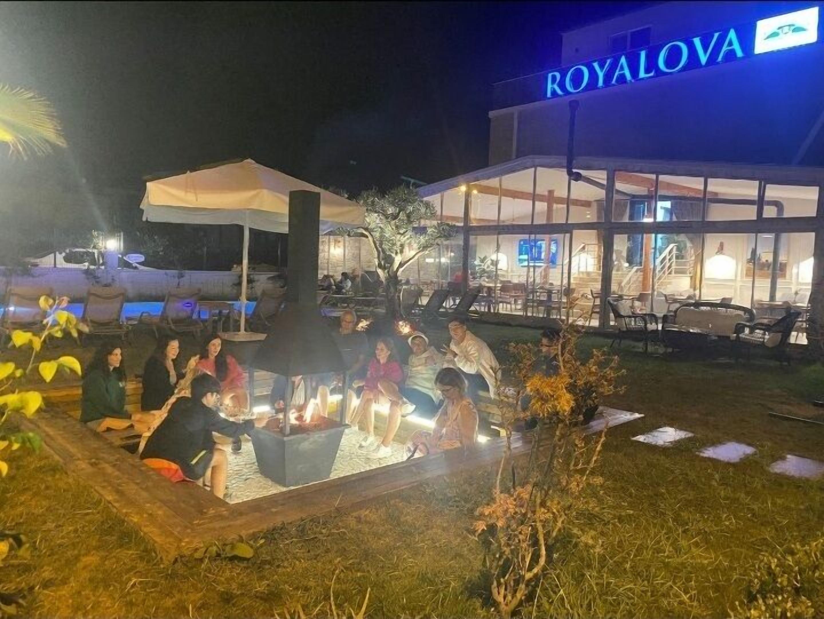 Un espace extérieur aménagé avec des sièges, un foyer et des tables, situé devant un bâtiment sur lequel on peut lire l’inscription « ROYALOVA ».