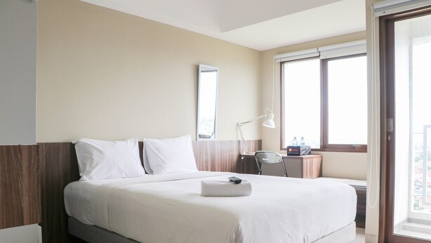 A hotel room with a large bed, a desk with a lamp, and a window with a city view. - Modern Look and Homey Studio Apartment LRT City Jatibening (Bekasi)