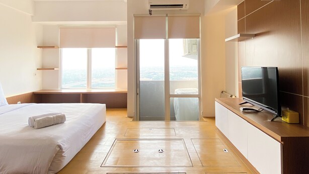 A modern hotel room with a bed, a TV, a sliding door to a balcony, and a view of the city. - Nice And Cozy Studio Apartment At Vasanta Innopark (Cikarang)