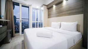 A hotel room with a large bed, a wooden headboard, and a city view through the window. - Homey Studio At Tamansari Papilio Apartment (Surabaya)