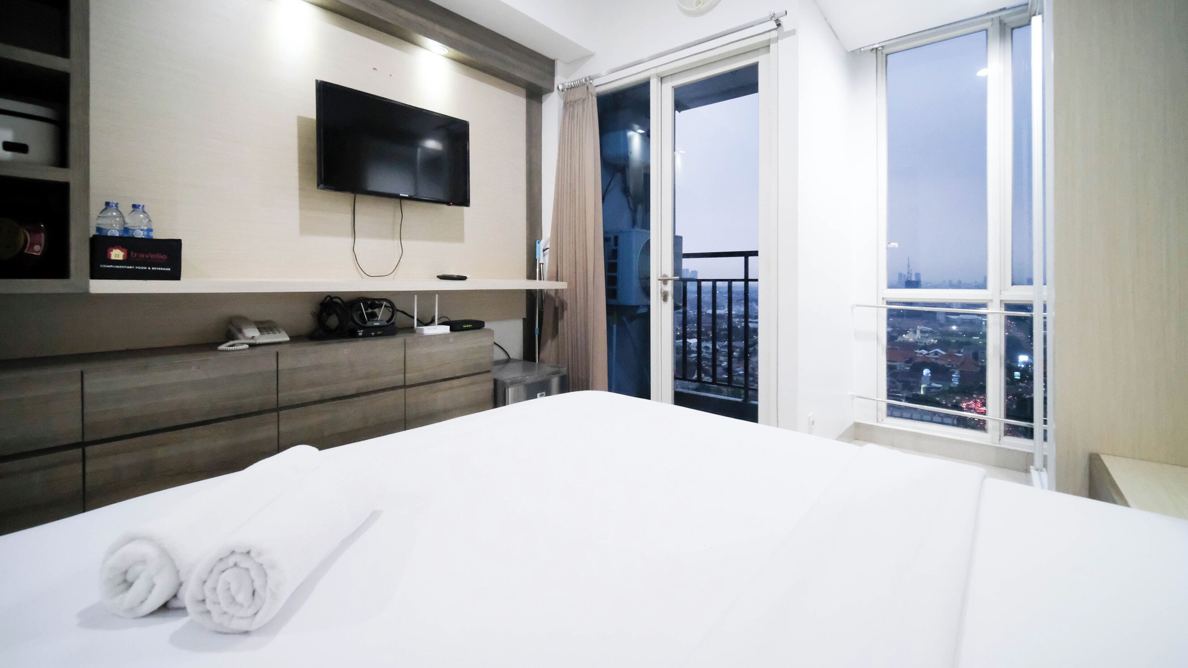 A hotel room with a large bed, a flat-screen TV mounted on the wall, a minibar, and a balcony with a city view.
