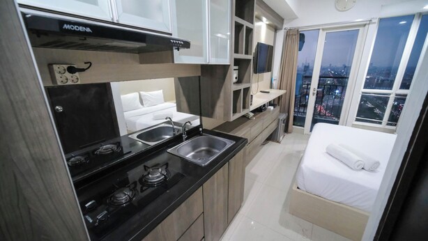 A compact kitchenette with a stove, sink, and microwave. A bed is visible with a city view through the window. - Homey Studio At Tamansari Papilio Apartment (Surabaya)