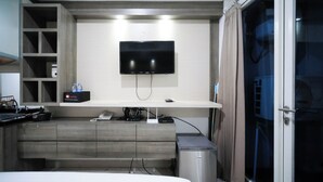 A modern hotel room with a flat-screen TV mounted on a white wall, a minibar, a desk with a telephone, and a GE appliance. - Homey Studio At Tamansari Papilio Apartment (Surabaya)
