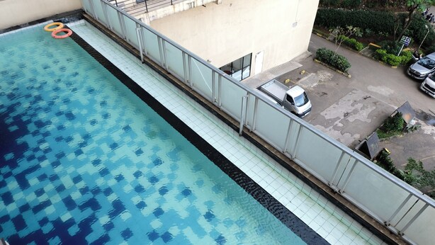 Una piscina con un diseño de mosaicos, una camioneta blanca estacionada cerca y un edificio con balcón que da a la piscina.