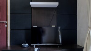 Una habitación moderna con una televisión de pantalla plana sobre un soporte de madera, una mesita blanca con un control remoto, y una puerta con cerradura.
