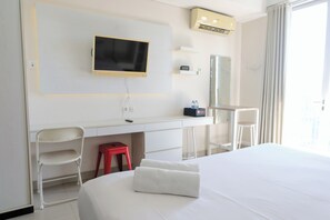 A hotel room with a bed, a desk, a chair, a TV, a mirror, and a window with curtains. - Homey And Cozy Studio Apartment At Dago Suites (Bandung)