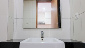 Een badkamer met een wit wastafelmeubel, een zwart aanrecht en een spiegel die een kamer met een bed en een deur weerkaatst.
