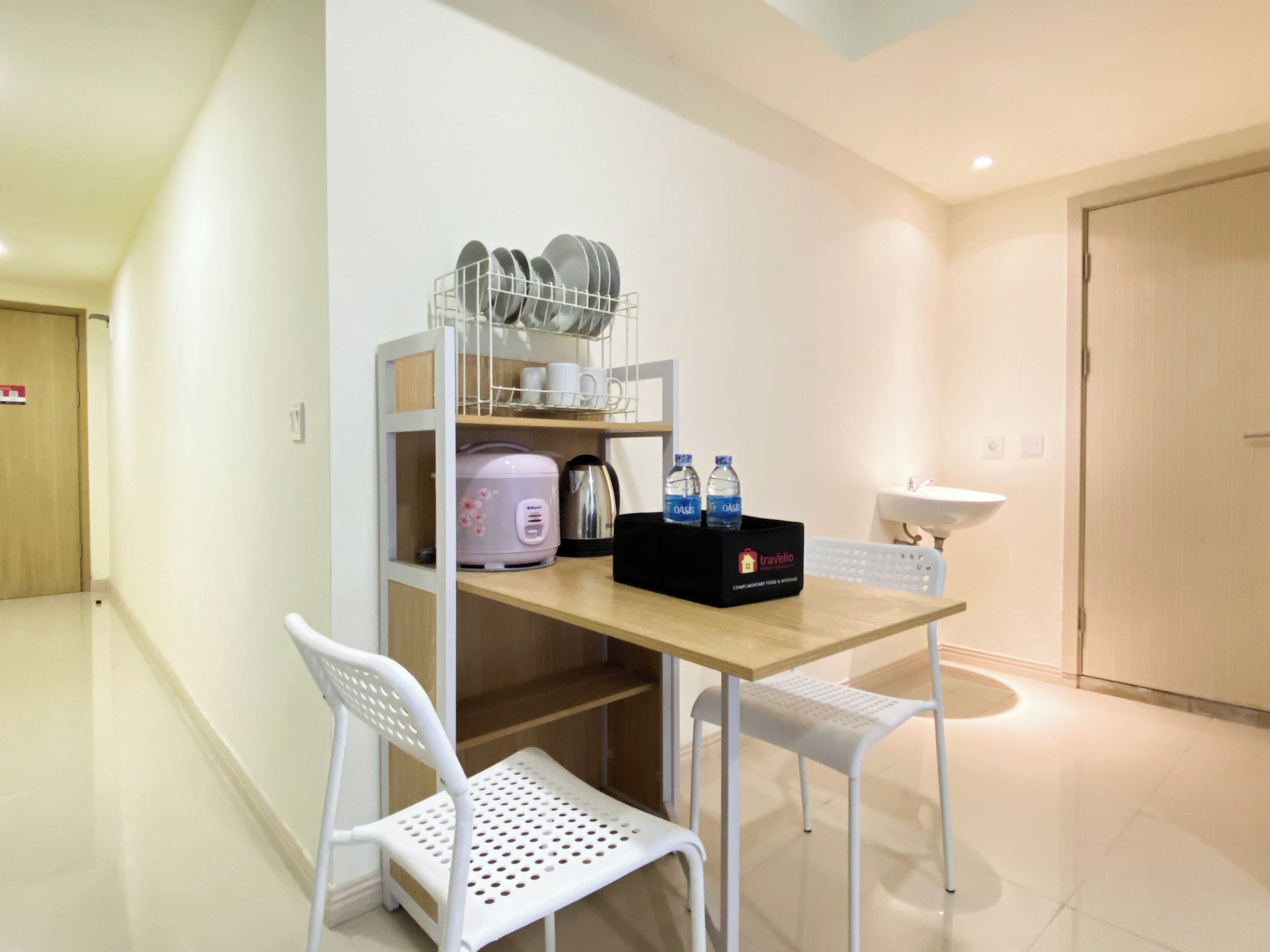A small kitchenette with a wooden table, two white chairs, a shelf with dishes, a pink appliance, and a box with water bottles.