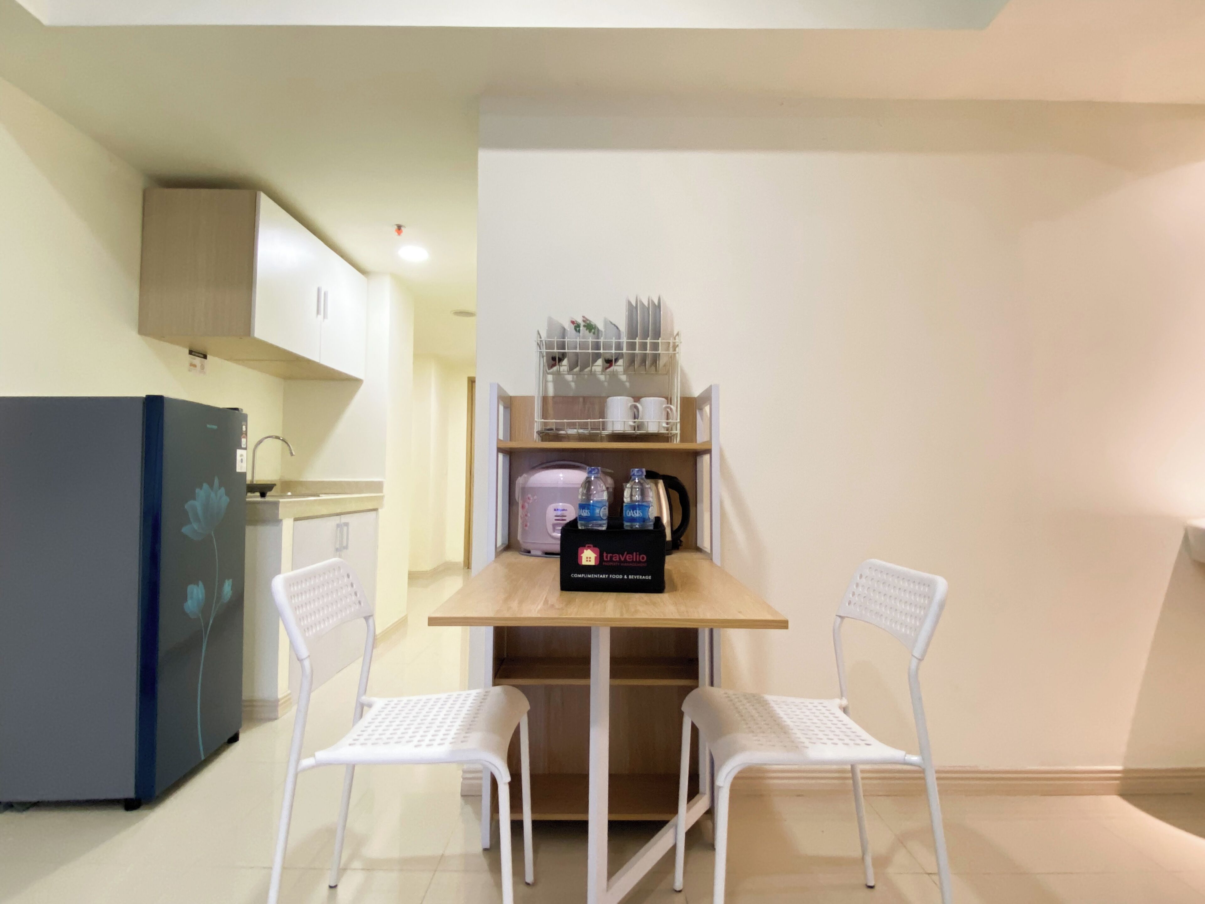 A compact kitchen with a refrigerator, a sink, and a countertop with a water dispenser and a box labeled "Travelio". There are two white chairs around a small table.